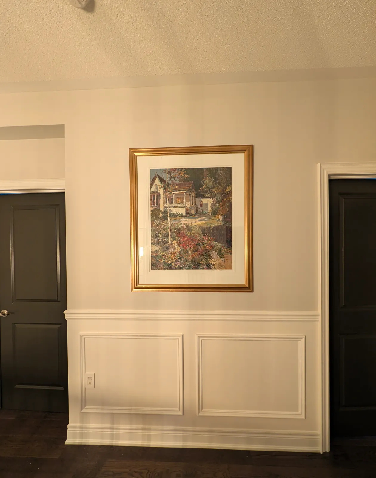 White chair rail wainscoting with two picture-frame panels on a hallway wall between two dark-stained doors, a gold-framed painting above