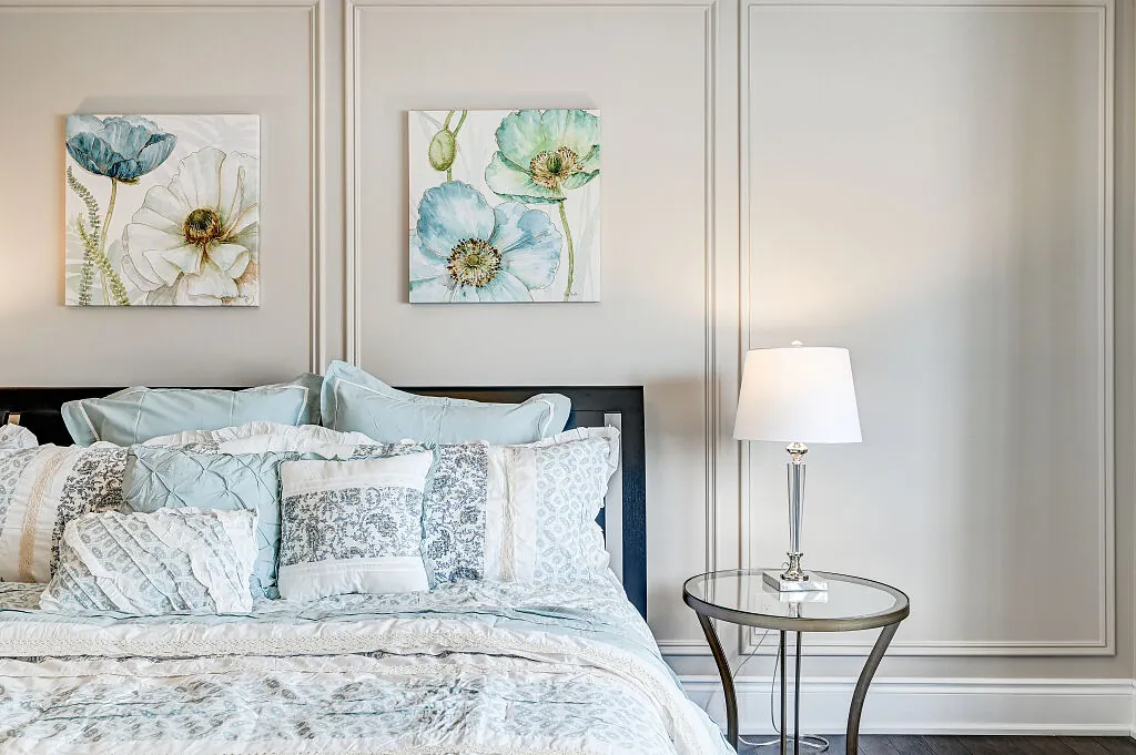 Staged bedroom with tall picture-frame wall panels behind the bed, two floral canvases and a glass side table with a lamp