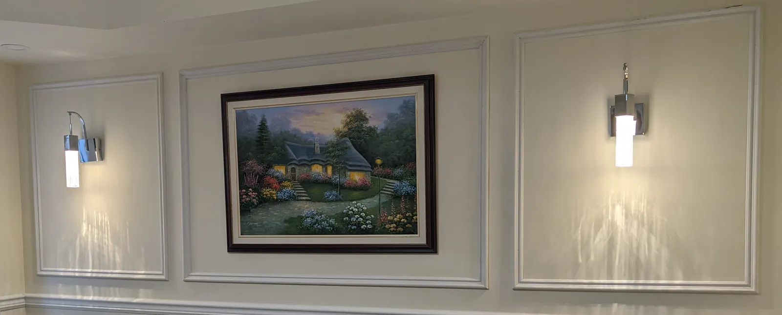 Dining room wall with picture-frame panels above a chair rail, two chrome wall sconces and a framed cottage painting