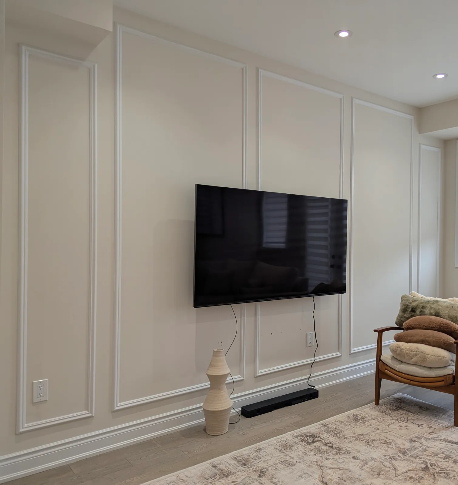 Cream living room wall with full-height picture frame panels around a wall-mounted TV, a wooden accent chair and a rug