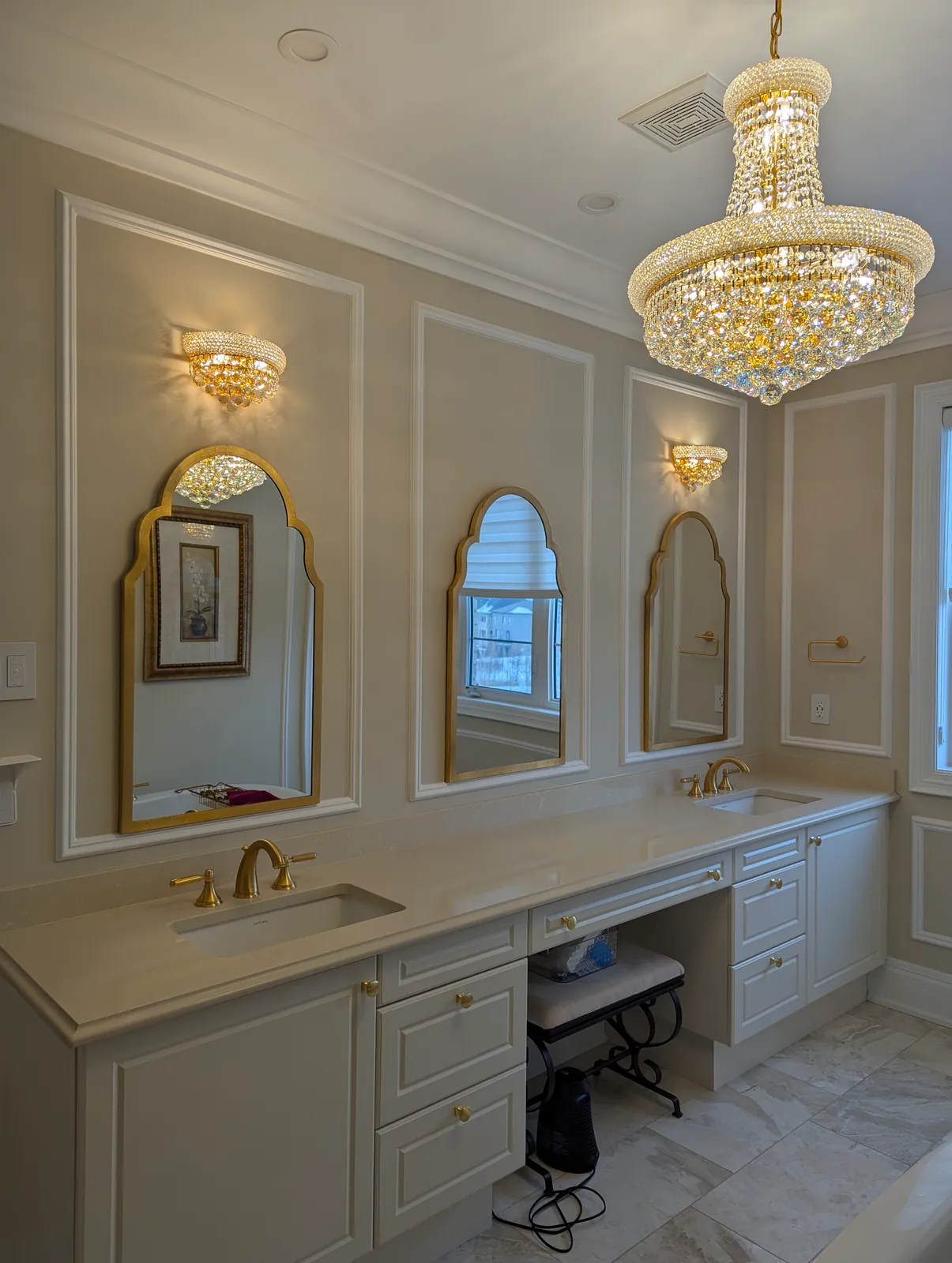 Bathroom with picture-frame panels framing three gold arched mirrors over a double vanity, crystal sconces and a chandelier
