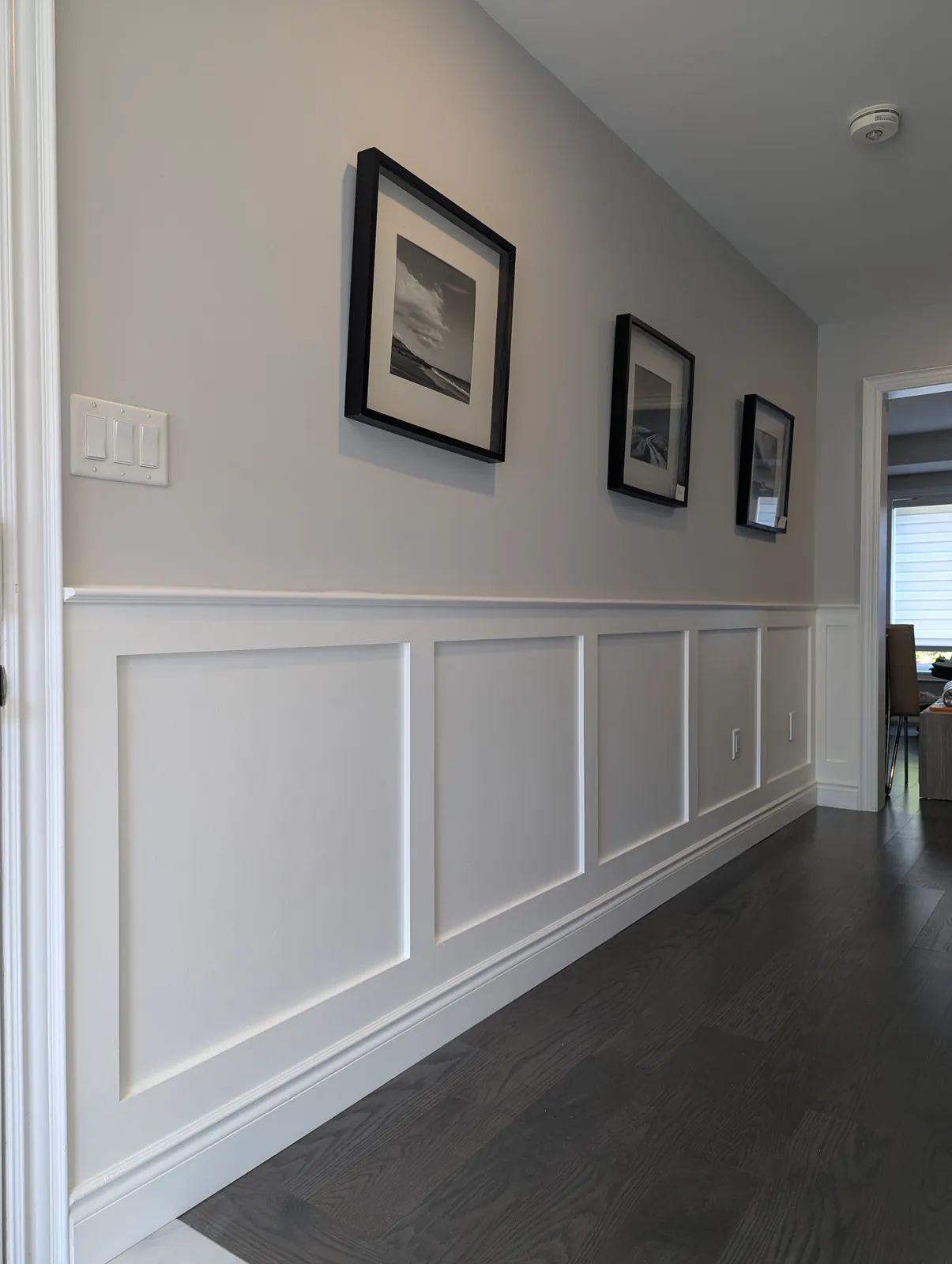 Hallway with white Shaker-style flat-panel wainscoting under a cap rail, grey walls with three black-framed photographs, dark hardwood floor